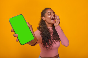 Young cheerful expressive Indian woman teenager with phone in hands shouts loudly announcing release of new mobile application or website for students stands in orange studio. Green screen