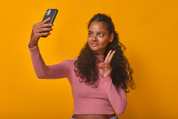 Young attractive smiling Indian woman with mobile phone in hands makes letter V with fingers and records video selfie on gadget to share important news with friends stands in orange studio.