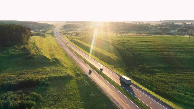 Truck driver with cargo is driving on intercity speed highway to customer through picturesque rural agricultural area with sunset background. Delivery and logistics of goods, aerial motion forward