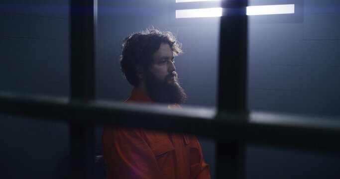 Believing male prisoner in orange uniform kneels near the bed, prays to God in prison cell holding Bible. Criminal serves imprisonment term for crime in jail or detention center. Faith in God concept.