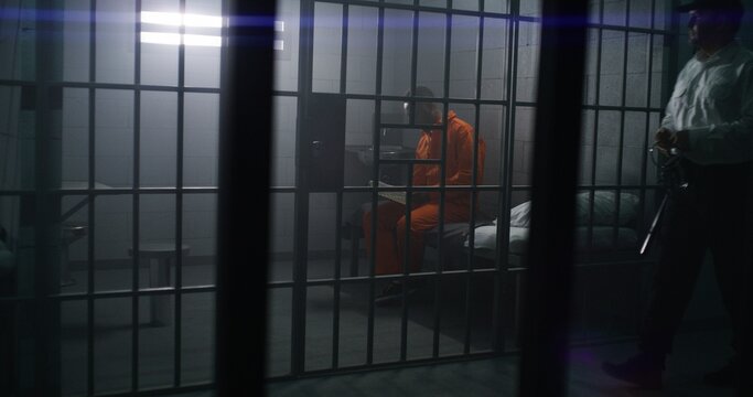 African American prisoner in orange uniform sits on the bed, reads Bible in prison cell. Prison officer walks, watches criminal. Inmate serves imprisonment term for crime in jail or detention center.