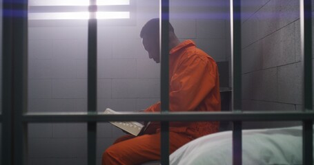 African American prisoner in orange uniform sits on bed behind bars, reads Bible in prison cell....