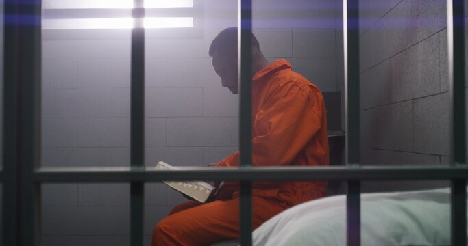 African American prisoner in orange uniform sits on bed behind bars, reads Bible in prison cell. Male criminal serves imprisonment term for crime in jail. Detention center or correctional facility.