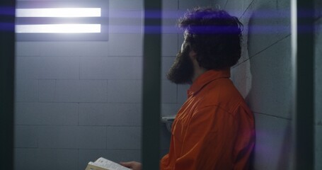 Male prisoner in orange uniform sits on the bed, reads Bible, looks at barred window in prison...