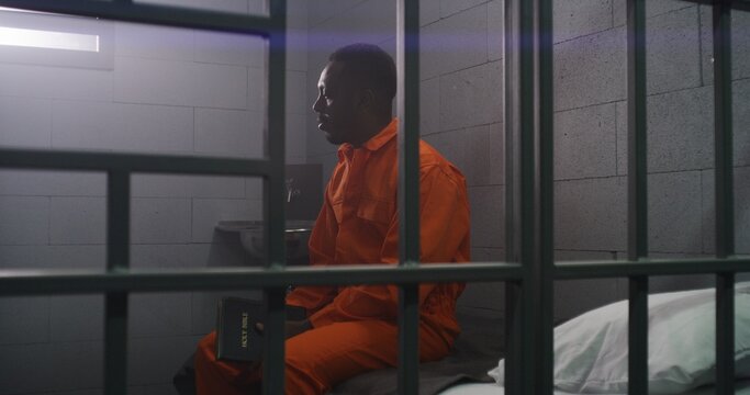 African American prisoner in orange uniform sits on the bed behind bars, reads Bible in prison cell. Criminal serves imprisonment term for crime in jail. Detention center or correctional facility.