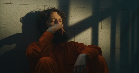 Depressed prisoner in orange uniform sits on bed, smokes cigarette in prison cell. Criminal serves...