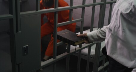 Prison officer gives Bible to male prisoner in orange uniform. Criminal sits on bed in prison cell, reads. Offender serves imprisonment term for crime in jail. Detention center. Faith in God concept.