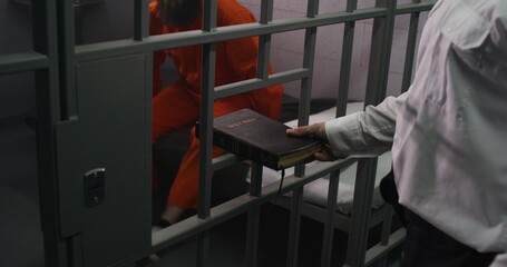 Prison officer gives Bible to male prisoner in orange uniform. Criminal sits on bed in prison cell,...