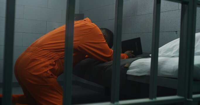Religious African American prisoner in orange uniform kneels near the bed, prays to God in prison cell holding Bible. Male criminal serves imprisonment term in jail or detention center. Faith in God.