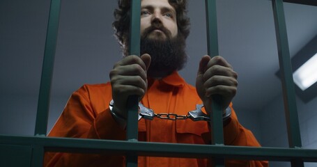 Male prisoner in orange uniform holds metal bars, stands in prison cell in handcuffs, looks at...