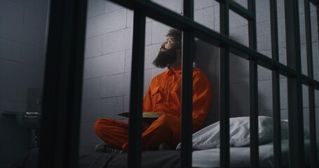 Male prisoner in orange uniform sits on bed, reads Bible, prays, looks at barred window in prison...