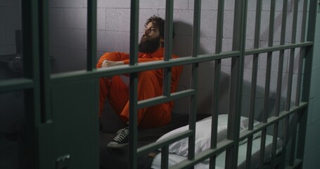 Male prisoner in orange uniform sits on bed, reads Bible, prays, looks at barred window in prison...