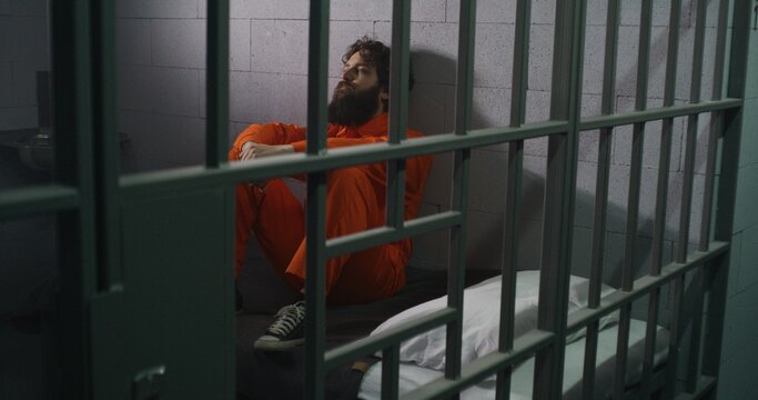 Male prisoner in orange uniform sits on bed, reads Bible, prays, looks at barred window in prison cell. Inmate serves imprisonment term for crime in jail. Correctional facility. Faith in God concept.