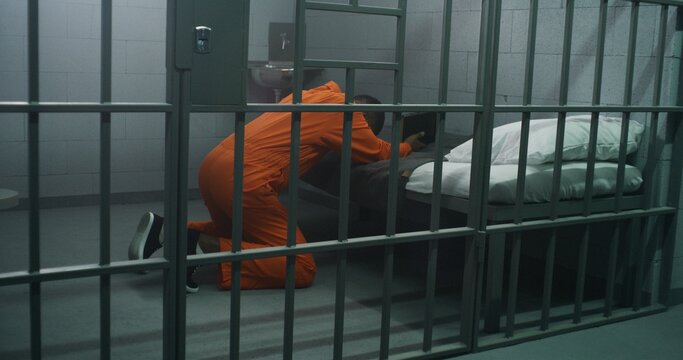 Religious African American prisoner in orange uniform kneels near the bed, prays to God in prison cell holding Bible. Male criminal serves imprisonment term in jail or detention center. Faith in God.