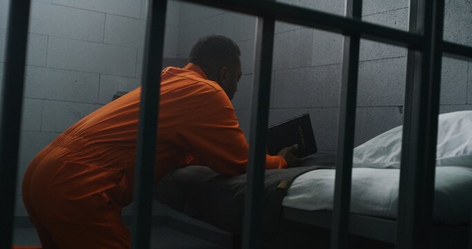 Religious African American prisoner in orange uniform kneels near the bed, prays to God in prison cell holding Bible. Male criminal serves imprisonment term in jail or detention center. Faith in God.