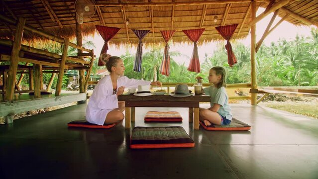 The happy family eat and drink tasty beverages spending time in local floating cafe on water. Mother and daughter having breakfast outdoors. Woman and a child in a cafe. Tropical view