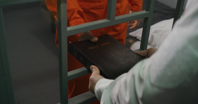 Prison officer gives Bible to male prisoner in orange uniform. Criminal sits on bed in prison cell, reads. Offender serves imprisonment term for crime in jail. Detention center. Faith in God concept.