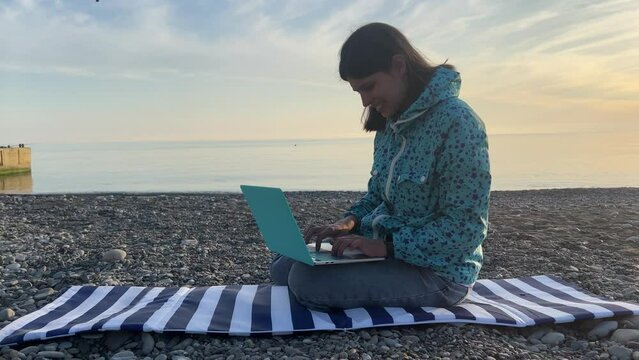 A young brunette sits on the beach by the sea and works typing on a laptop. Side view. The concept of remote work. High quality 4k footage