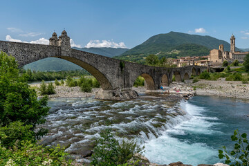 The ancient Ponte Gobbo over the Trebbia river with Bobbio, Italy in the background on a sunny summer day - obrazy, fototapety, plakaty