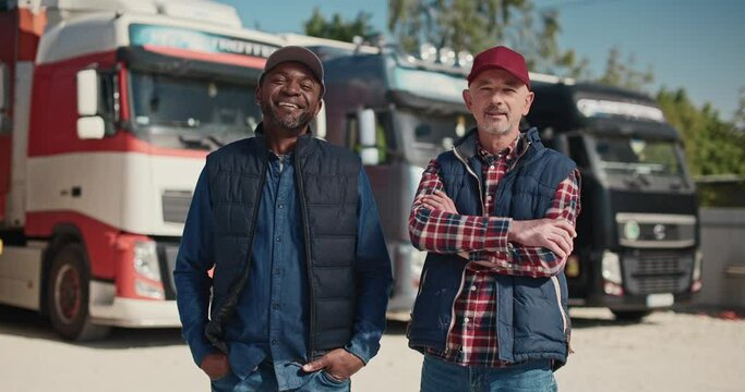 Two mixed-race coworkers standing before large trucks outside. Mature drivers in hats and jackets positively looking into camera. Getting ready to traveling. Working together in delivery company.