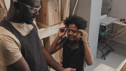 Tilt up shot of dad putting on glasses on son when preparing for safe crafting in carpentry workshop