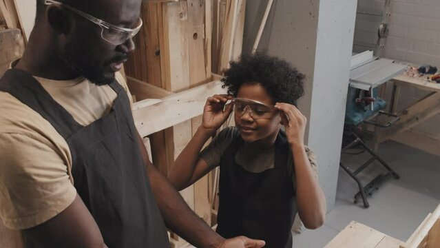 Tilt up shot of dad putting on glasses on son when preparing for safe crafting in carpentry workshop