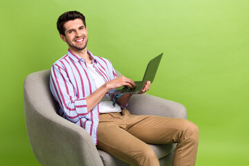 Photo of good mood man with stubble dressed striped shirt typing email on laptop sit in armchair isolated on green color background