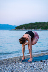 A young girl in a black tank top and pink shorts doing yoga on the sea beach. Yoga at sunset overlooking the beautiful Adriatic Sea and green shores. Photo promotes a healthy lifestyle, sports, yoga.