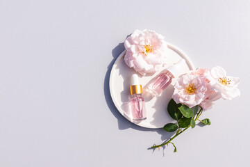 Two transparent cosmetic bottles with a pipette with natural rose oil or serum based on rose petal oil on a white plate and flowers. Top view.