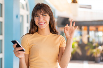 Young woman at outdoors using mobile phone and doing OK sign