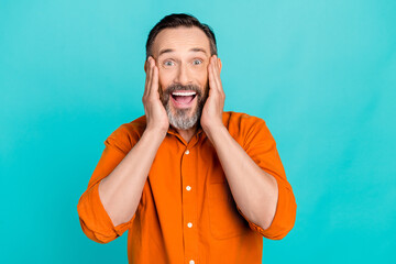 Photo of shocked excited man dressed orange shirt arms cheeks open mouth isolated turquoise color background