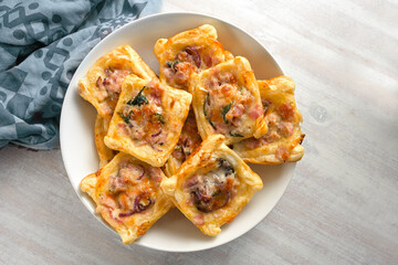 Baked puff pastry squares with ham, onion, herbs and cheese in a bowl, finger food snack for a warm or cold party buffet, high angle view from above, copy space, selected focus