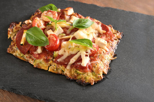 Piece of low carb pizza on a crust made from zucchini and oat flakes, topped with tomato, feta cheese and salami on a dark slate plate, copy space, selected focus
