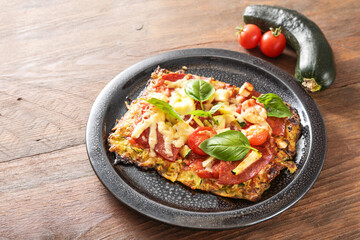 Piece of low carb pizza on a crust from zucchini and oat flakes, topped with tomato, feta cheese, salami and basil, dark plate on a wooden table, copy space, selected focus