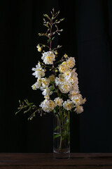 Vintage rose branch with yellow white flowers in a glass vase against a dark background, copy space, vertical