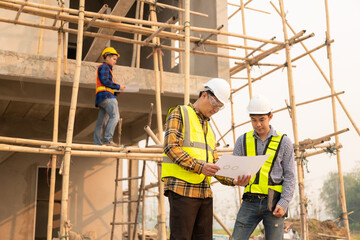 Architects and engineers working together sketching building plans and discussing at construction site wearing uniforms and helmets standing on construction site about large industrial architecture.