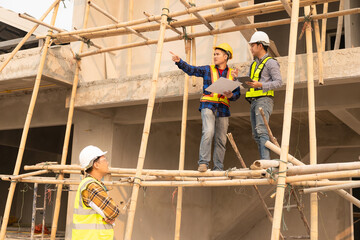 Architects and engineers working together sketching building plans and discussing at construction site wearing uniforms and helmets standing on construction site about large industrial architecture.