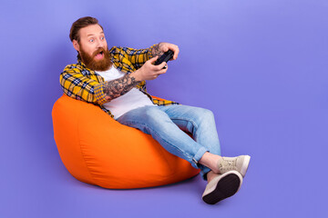 Photo of cheerful guy man wear stylish clothes sit comfy pouf have fun playstation watch empty space isolated on purple color background