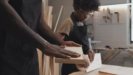 African American father and son working with surface of wooden plank while standing next to table together in carpentry studio