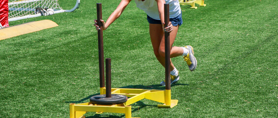 Young female pushing a yellow sled with wight on a turf field