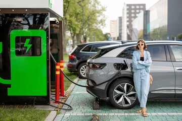 Young woman waiting for her electric car to be charged on a public charging station at city. Concept of modern lifestyle and green energy for transportation