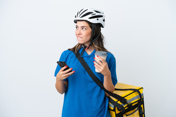 Young delivery woman with thermal backpack isolated on white background holding coffee to take away and a mobile while thinking something