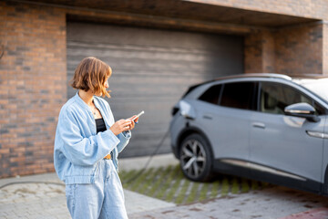 Young woman useing smart phone while charging her electric car near garage of her house. Concept of...