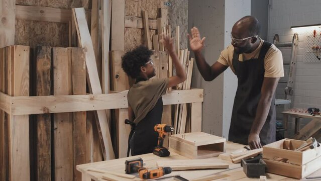 African American positive dad giving five to his son during crafting on table in carpentry workshop