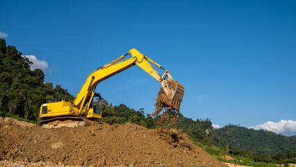 excavator machine is at work digging and dumping soil above the hills with tropical forest in the...