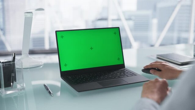 Over the Shoulder Footage with Corporate Employee Using Laptop Computer with Mock Up Green Screen Chromakey Display. Man Sitting Behind a Desk, Browsing Internet, Using a Wireless Mouse