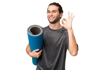 Young sport man going to yoga classes while holding a mat over isolated background showing ok sign with fingers © luismolinero