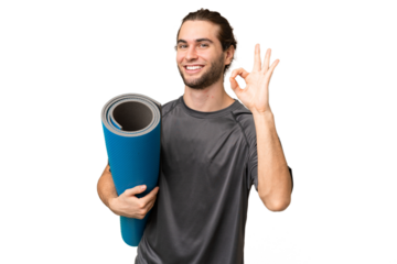 Young sport man going to yoga classes while holding a mat over isolated background showing ok sign with fingers