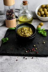 pesto sauce in a black bowl with olives, olive oil and basil. stone background. 
