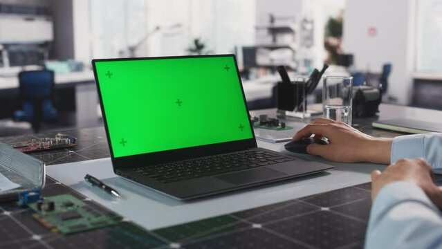 Over the Shoulder Footage with Industrial Engineer Using Laptop Computer with Mock Up Green Screen Chromakey Display. Specialist Sitting Behind a Desk, Browsing Blueprints and Project Plans